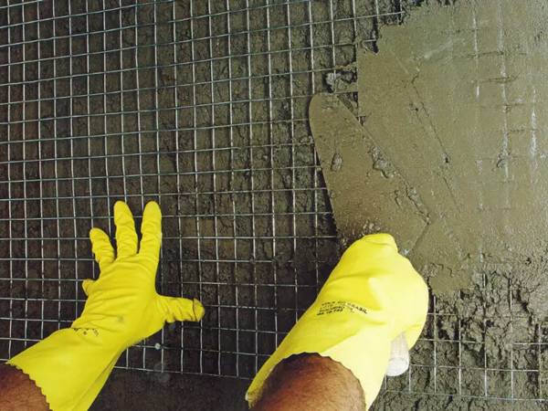 A man with yellow gloves is plastering the wall with welded wire mesh.